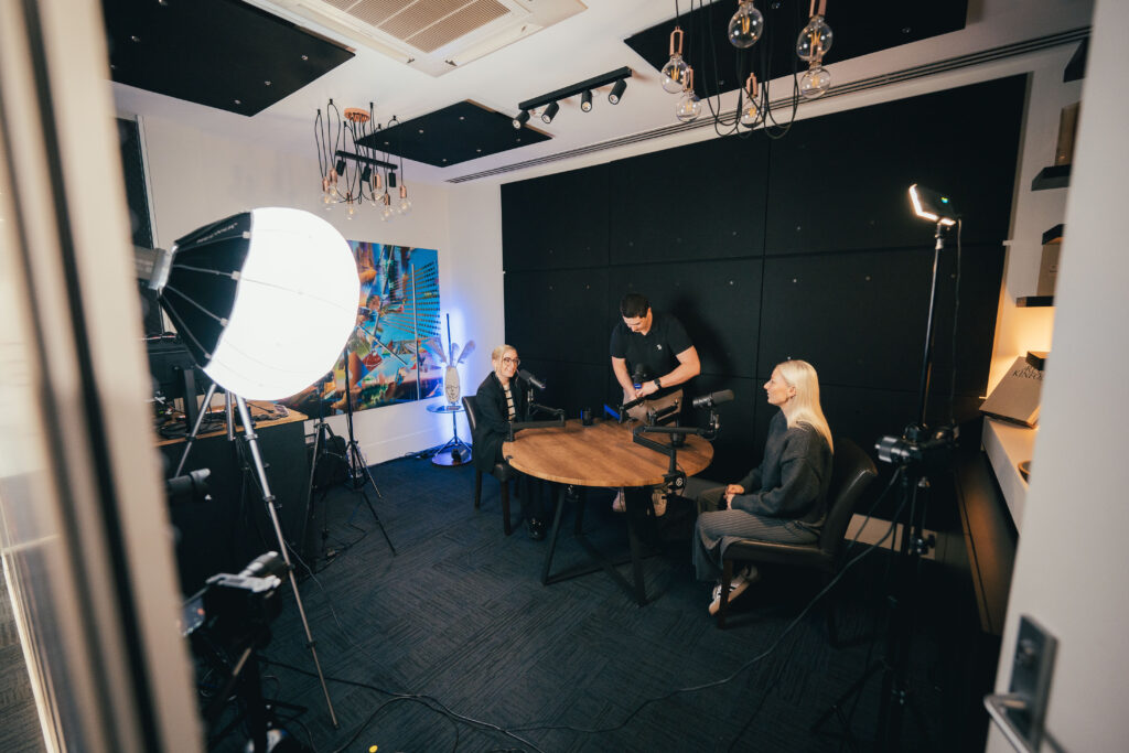 Wide angle view of Podwave Studios West Perth with producer setting up cameras while two guests wait at a round table with white walls and Edison lighting