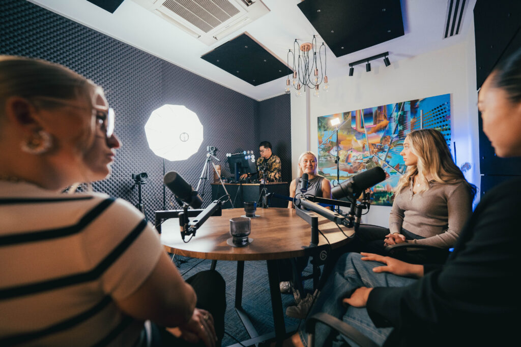 Three women in conversation at microphones recording a group podcast at Podwave Studios West Perth with producer visible in background at monitor