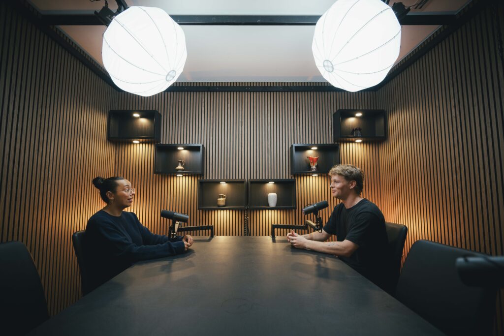 Wide shot of Talietha and Christian in conversation at microphones in the full West Leederville studio with overhead ring lights and timber panelling